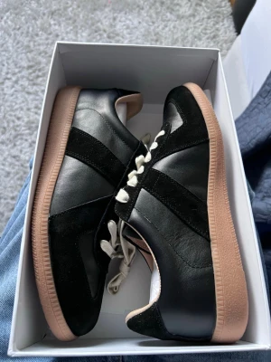 Maison Margiela gats - Stilrena svarta sneakers med beige gummisula och vita snören. Skorna har en mix av läder och mockadetaljer som ger en schysst kontrast. Perfekta för dig som gillar en clean och klassisk look med lite retrovibbar.