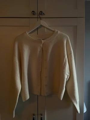 Krämvit kofta med knappar - Mysig krämvit kofta med rund halsringning och knäppning framtill. Koftan har långa ärmar, ribbade muddar och en något boxig, croppad passform. Aldrig använd och är köpt på Missay i Göteborg.(orginalpris 500)