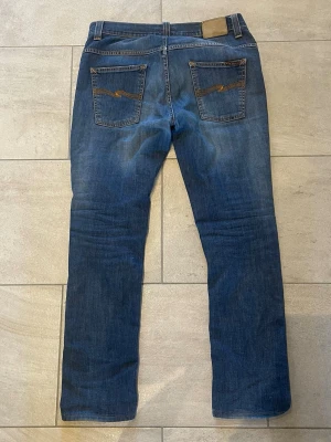 Mörkblå nudie jeans - Snygga mörkblå jeans med lätt slitning och klassisk femficksdesign. Jeansen har normal midja och raka ben. Perfekta för en avslappnad och trendig look. Jeans är i modellen thinn finn och passar någon som bär 32/32 i storlek