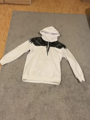 Vit hoodie med svart vingtryck - Snygg vit hoodie med svart vingmotiv över axlar och bröst, samt huva och magficka. Trycket fortsätter även på ryggen för en cool look. Perfekt för dig som vill sticka ut med en unik design. Mjuk och skön bomullskänsla. Passar M och L. (Marcelo burlon heter märket)
