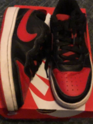 Nike Air Jordan 1 Low svart/röd - Säljer ett par Nike Air Jordan 1 Low sneakers i svart och rött med klassisk röd swoosh och vit sula. Skorna har perforerad tå, svarta skosnören och den ikoniska Air Jordan-designen. Perfekta för dig som gillar streetwear och sportig stil.