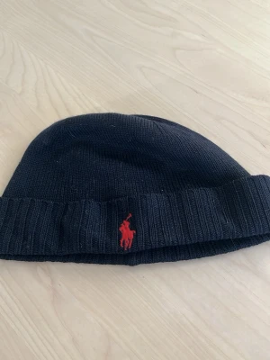 Mörkblå mössa  från Polo Ralph Lauren - Säljer en mörkblå ribbstickad mössa från Polo Ralph Lauren med den klassiska röda logotypen broderad framtill. Mössan är mjuk och stretchig, perfekt för kyliga dagar. Enkel och stilren design som passar till det mesta. Pris är diskuterbart!