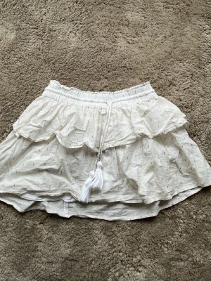 Vit volangkjol med tofsdetaljer - Supersöt kort vit kjol med volanger och små guldfärgade prickar. Kjolen har resår i midjan och dekorativa tofsar med pärlor framtill. Perfekt för sommarens alla tillfällen och ger en romantisk vibe. Är i barn stolek och har används fåtal gånger! Kjolen är vitare påriktigt!