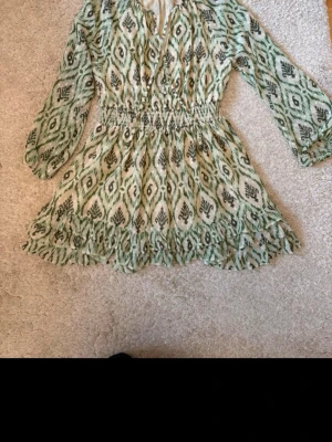 Grön mönstrad kortklänning med volang - Snygg kortklänning med grönt och vitt mönster, långa ärmar och smock i midjan. Klänningen har volangdetaljer nertill och är tillverkad i ett lätt, luftigt material. Perfekt för vår och sommar!