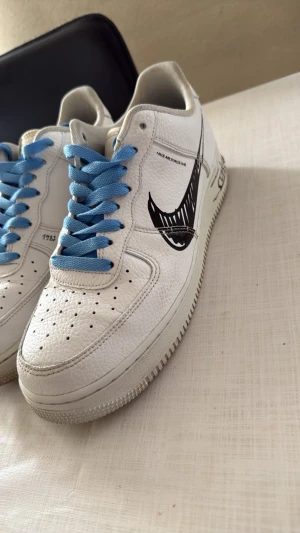 Nike Air Force 1 med blå snören - Säljer ett par vita Nike Air Force 1 sneakers i läder med unika svarta detaljer på sidan och blå skosnören. Klassisk perforerad tå och låg modell. Perfekta för dig som vill sticka ut med en personlig touch på dina sneakers.