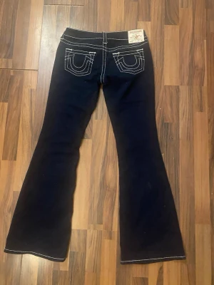 Mörkblå låg midja bootcut jeans från True Religion - Säljer ett par svarta bootcut jeans från True Religion med tydliga vita kontrastsömmar. Klassisk femficksmodell och ikoniska broderade bakfickor. Jeansen har låg midja och är tillverkade i denim med stretch för extra komfort. Orginal pris 1 399kr