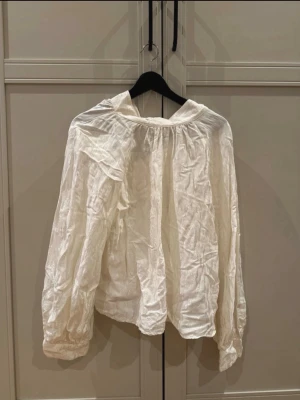 Vit luftig blus med lång ärm - En vit, luftig blus med  halsringning och långa ärmar med breda muddar. Blusen har en avslappnad passform och är tillverkad i ett lätt, något glansigt tyg som ger en trendig och stilren look.