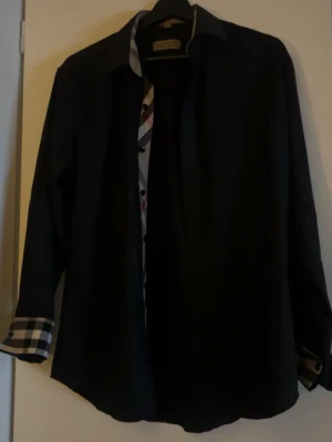 Svart skjorta med rutiga detaljer - Stilren svart skjorta från Burberry med klassiska rutiga detaljer på insidan av kragen och manschetterna. Skjortan har lång ärm och knappar framtill. Inga märken eller så, säljer den för att den inte passar