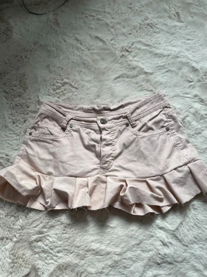 Ljusrosa volangkjol med fickor - Supersöt ljusrosa kortkjol med volangkant och klassiska jeansfickor både fram och bak. Kjolen har knapp och dragkedja framtill och är gjord i mjuk bomull. Perfekt för dig som gillar en fem och avslappnad stil