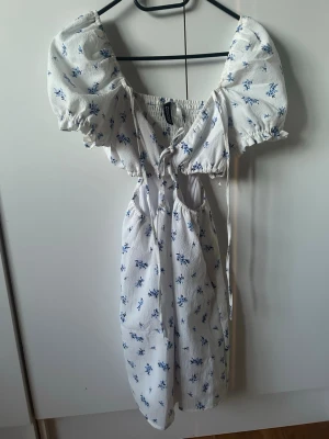 Vit blommig kortklänning med puffärm - Supersöt vit kortklänning med blått blommönster och puffärmar. Klänningen har omlottdetalj med knyt framtill, smock i ryggen och en lätt volangkant vid ärmslut. Perfekt för sommarens alla tillfällen!