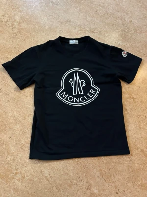 Svart Moncler t-shirt med stor logga - Snygg svart t-shirt från Moncler med stor vit logga på bröstet och klassisk rund hals. T-shirten har korta ärmar och en liten Moncler-patch på ena ärmen. Tillverkad i mjuk bomull. Ny fast utan lapparna kvar. Storlek small passar någon som är XS