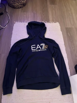 Mörkblå hoodie från Emporio Armani - Snygg mörkblå hoodie från Emporio Armani EA7 med stor logga på bröstet och klassisk huva. Tröjan har en stor magficka och ribbade muddar vid ärmslut och nederkant. Perfekt för dig som vill ha en stilren och trendig look. Står ej något om någon storlek men skulle tippa på att det är S då den passar mig som är 160. Använd ett fåtal gånger men inget som syns.