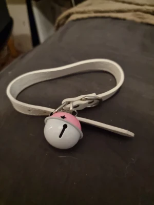 Vitt choker med bjällra och rosa detalj - Snyggt vitt choker i syntetmaterial med en  bjällra som har rosa och vit detalj samt stjärnformad utskärning. Perfekt accessoar för att ge din outfit en lekfull touch. Justerbar längd med klassiskt spänne.