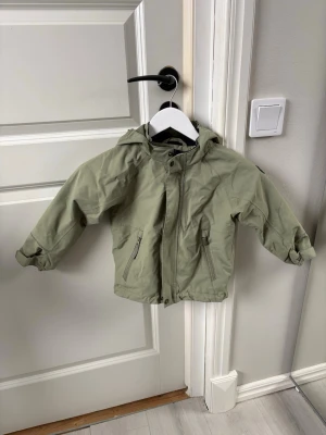 Grön vindjacka från Polarn O. Pyret - Info står på bild 3. Flexi size = kan ändra storlek vid midjan och ärmen med hjälp av inbyggda band. Liten fläck som knapp syns på ena ärmen, syns på bild 4