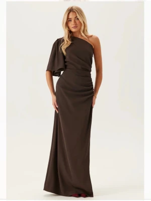 Brun balklänning  - En väldigt fin och elegant brun balklänning från Bubbleroom med oneshoulder-design och draperad detalj i midjan. Klänningen har en diskret dragkedja vid midjan och en slits vid sidan. Helt oanvänd med prislappen kvar. Slutsåld på hemsidan!