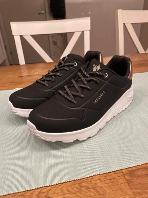 Svarta Skechers sneakers med vit sula - Svarta sneakers från Skechers med chunky vit sula och detaljer i guld vid hälen. Skorna har perforerad ovandel, svarta och vita snören samt Skechers-logga på sidan. Perfekta för en sportig och stilren look.