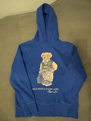 Blå hoodie Polo Ralph Lauren - Blå hoodie från Polo Ralph Lauren med ikoniskt Polo Bear-tryck på bröstet. Klassisk huva och ribbade muddar. Mjuk bomullskänsla och avslappnad passform, perfekt för en cool och bekväm stil.