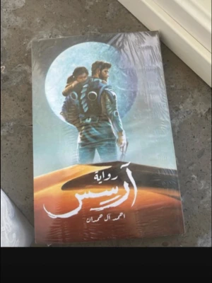 Science fiction-roman på arabiska - Säljer en skönlitterär bok med science fiction-tema på arabiska. Omslaget visar två personer i futuristiska dräkter framför en stor måne och ökenlandskap. Perfekt för dig som gillar äventyr och framtidsberättelser.