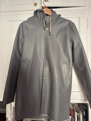 Grå regnjacka från Stutterheim - Snygg grå regnjacka från Stutterheim med minimalistisk design, huva med dragsko och tryckknappar framtill. Missfärgningar inuti jackan, synd ingenting utanpå/när man bär den. Storlek XS men jag är vanligtvis M. Funkar för många beroende på hur stor man vill ha den! 