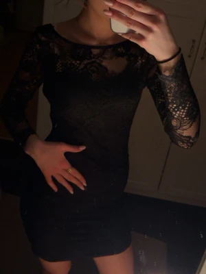 Svart spetsklänning med lång ärm - Säljer en svart kortklänning med långärmade spetsdetaljer och rund halsringning. Klänningen har ett elegant spetsmönster över hela och är figurnära med transparenta ärmar. Perfekt för dig som vill ha en stilren och snygg look.