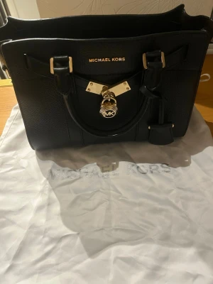 Svart Michael Kors handväska i läder - Snygg svart handväska från Michael Kors i strukturerat läder med guldfärgade metalldetaljer och lås framtill. Väskan har dubbla handtag och klassisk rektangulär form. Perfekt för dig som vill ha en stilren och lyxig accessoar. Äkta 