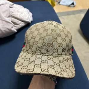 Beige keps med monogrammönster - Snygg beige keps från Gucci med det ikoniska monogrammönstret. Kepsen har gröna och röda ränder på sidan och justerbart spänne bak. Perfekt för att ge din outfit en lyxig touch. Pris kan diskuteras