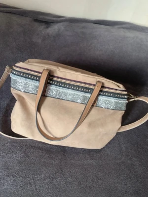 Beige handväska med grått band - Stilren och rymlig väska i mockaimitation med dekorativa detaljer i silverglitter och nitar✨. Väskan har både korta handtag och en justerbar, avtagbar axelrem vilket gör den flexibel att bära både över axeln och i handen.🩶 Den har även fickor på insidan och ut 