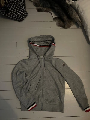 Grå Moncler hoodie med dragkedja - Snygg grå hoodie från Moncler med dragkedja framtill och huva. Detaljer i rött, vitt och blått på muddar och huvkant samt Moncler-logga på ärmen. Tillverkad i mjuk bomullsblandning för skön känsla. Perfekt för en avslappnad och stilren look.