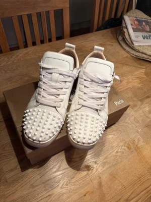 Vita Christian Louboutin sneakers - Säljer ett par vita sneakers från Christian Louboutin med ikoniska röda sulor och coola nitar över hela tån. Skorna har lågt skaft, vita skosnören och är tillverkade i läder. Perfekta för dig som vill sticka ut med en lyxig och edgy stil.