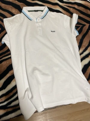 Vit pikétröja från BOSS - Snygg vit pikétröja från BOSS med klassisk krage och knäppning framtill. Kragen har blå och svarta ränder som ger en fräsch touch. Broderad BOSS-logga på bröstet. Perfekt för en stilren och sportig look. Varför den saknar en tyg lapp är för att den störde och kliade mig mycket när jag hade på mig den men det står på den att det är storlek 4xl men boss kläder är ju mindre i storleken samt att jag har tagit den till en skräddare och gjort den mindre så det är storlek XL nu på den. 