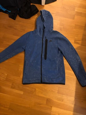 Blå Nike hoodie med dragkedja - Snygg blå hoodie från Nike med hel dragkedja och huva. Tröjan har en svart bröstficka med dragkedja och två sidofickor. Materialet ser ut att vara mjukt och sportigt, perfekt för en avslappnad stil. Nike-loggan finns på bröstet.