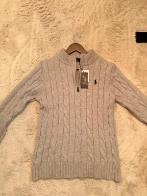 Beige kabelstickad tröja Polo Ralph Lauren - Kabelstickad ljusgrå tröja från Polo Ralph Lauren med halv dragkedja och hög krage. Säljer den för den är för liten på mig! (Är 185cm) Skule säga att den passar personer runt 175-180. Så skulle säga att den är mer som en storlek M än L. Längd: 64cm