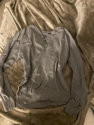 Grå v-ringad tröja med knappar - Grå långärmad tröja med v-ringning och dekorativa knappar längs halsringningen. Tröjan har ribbade muddar vid ärmslut och nederkant samt en diskret broderad logga på bröstet. Perfekt för en avslappnad och stilren look.