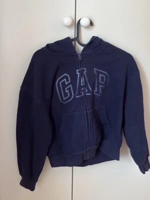 Mörkblå hoodie med dragkedja från GAP - Mörkblå cropped hoodie från GAP med dragkedja. Hoodien är använd men i gott skick. Storlek Kids XL 12 år.