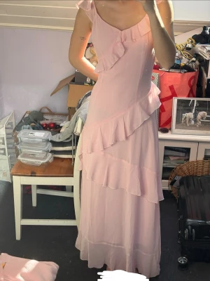 Ljusrosa långklänning med volanger - Söt ljusrosa långklänning med volangdetaljer från NA-KD, nypris 700kr. Klänningen har en slits på ena sidan samt dragkedja och är djup i ryggen. Jag är 170 cm för ländreferens. Använd en gång så är i jättebra skick, säljer för att den är för stor för mig (håller in den på första bilden). Perfekt till balen!💕