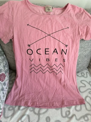 Rosa t-shirt med tryck från Tribute strl: M - Säljer en rosa t-shirt från Tribute med svart tryck 'Ocean Vibes' och grafiska pilar och vågor. T-shirten har rund halsringning och korta ärmar. Perfekt för en avslappnad stil och tillverkad i mjuk bomull.