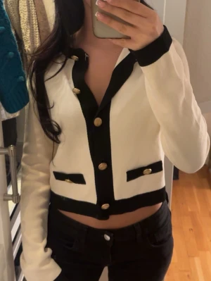 Vit och svart cardigan med guldknappar - Trendig vit cardigan med svarta detaljer och dekorativa guldknappar framtill. Kort modell med två fejkfickor och markerad krage. Perfekt för en chic och stilren look.