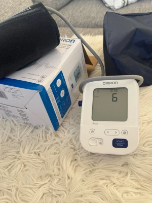 Omron blodtrycksmätare digital - Digital blodtrycksmätare från Omron med stor display och tydliga knappar. Vit och blå design med svart manschett och slang. Kommer med originalkartong och förvaringspåse. Perfekt för att enkelt hålla koll på blodtrycket hemma.