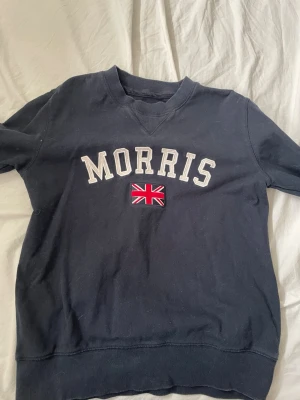 Mörkblå stickad tröja Morris - Mörkblå stickad tröja från Morris med broderad text och brittisk flagga på bröstet. Tröjan har rund halsringning och ribbade muddar vid ärmslut och nederkant. Perfekt för en stilren och avslappnad look.