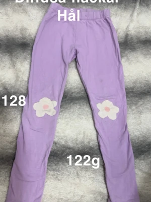 Lila leggings med blommor på knäna - Säljer ett par lila leggings med vita blommor och rosa mitt på knäna. De har resår i midjan. Perfekta för en färgglad och lekfull stil.🌸 Hål i rumpan. Diffusa fläckar. Storlek 128 