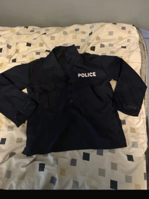 Polisjacka för maskerad - Mörkblå polisjacka med vita bokstäver 'POLICE' på bröstet. Jackan har knäppning framtill och två fickor. Perfekt för maskerad eller cosplay när du vill klä ut dig till polis.