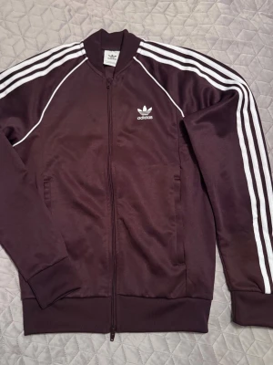 Adidas adicolor jacka zip tröja - En adidas originals adicolor tröja med dragkedja.  Storlek : S ,unisex ,smal passform , fit slim. Skulle säga att det är smal passform.  Färg : blandning mellan vinröd och mörklila, plommon färgad .  Tröjan är endast testad ! Inga defekter !