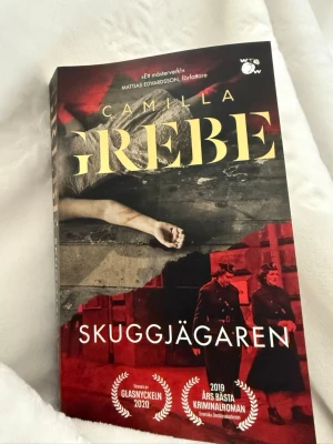 Skuggjägaren av Camilla Grebe - Säljer boken Skuggjägaren av Camilla Grebe, en spännande kriminalroman i pocketformat med färgstark omslagsdesign i svart, rött och gult. Boken är tryckt på papper och har prisbelönats med Glasnyckeln 2020 och Årets bästa kriminalroman 2019.