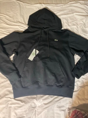 Svart hoodie från Lacoste - Snygg svart hoodie från Lacoste med klassisk krokodillogga på bröstet. Hoodien har huva med dragsko, stor magficka och ribbade muddar vid ärmar och nederkant. Perfekt för en chill och stilren look.