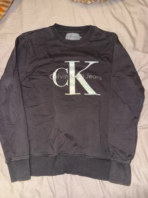 Svart sweatshirt från Calvin Klein - Snygg svart sweatshirt från Calvin Klein Jeans med stort CK-logotyptryck på bröstet. Tröjan har rund halsringning, långa ärmar och ribbade muddar vid ärmslut och nederkant. Perfekt för en avslappnad och trendig look.