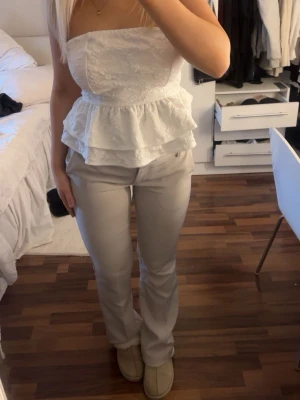 Vit axelbandslös volangtopp - Säljer en söt vit axelbandslös topp med volanger och spetsdetaljer. Toppen har en bandeauformad överdel och knyts med en stor rosett i ryggen. Perfekt för sommarens alla tillfällen!