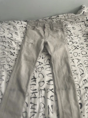 Ljusgrå jeans från Dsquared2 - Säljer ett par ljusgrå  från Dsquared2 med klassisk femficksdesign och en liten sliten detalj på vänster lår. Jeansen har en diskret röd logotagg vid fickan och är tillverkade i mjukt denimtyg. Perfekta för en trendig och avslappnad look.