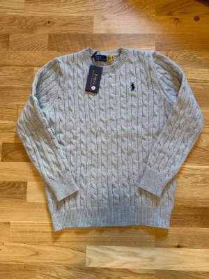 Polo Ralph Lauren - Ljusgrå kabelstickad tröja från Polo Ralph Lauren med rund halsringning och broderad logga på bröstet. Tröjan har ribbade muddar och är tillverkad i mjukt garn för en klassisk och stilren look.