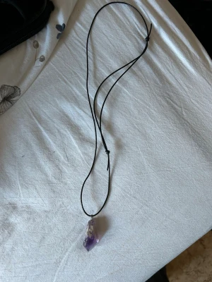Halsband med lila kristallhänge - Snyggt halsband med svart snöre och ett spetsigt hänge av lila kristall ametist. Enkelt och stilrent, perfekt för dig som gillar kristaller och vill ha en unik accessoar. Ingen synlig märkning på halsbandet.