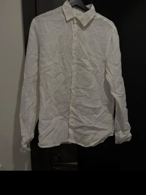 Vit linneskjorta från Dressmann - Vit skjorta från Dressmann i 100% linne. Skjortan har klassisk krage, knäppning framtill med vita knappar och långa ärmar. Perfekt för en stilren och luftig look under varma dagar.